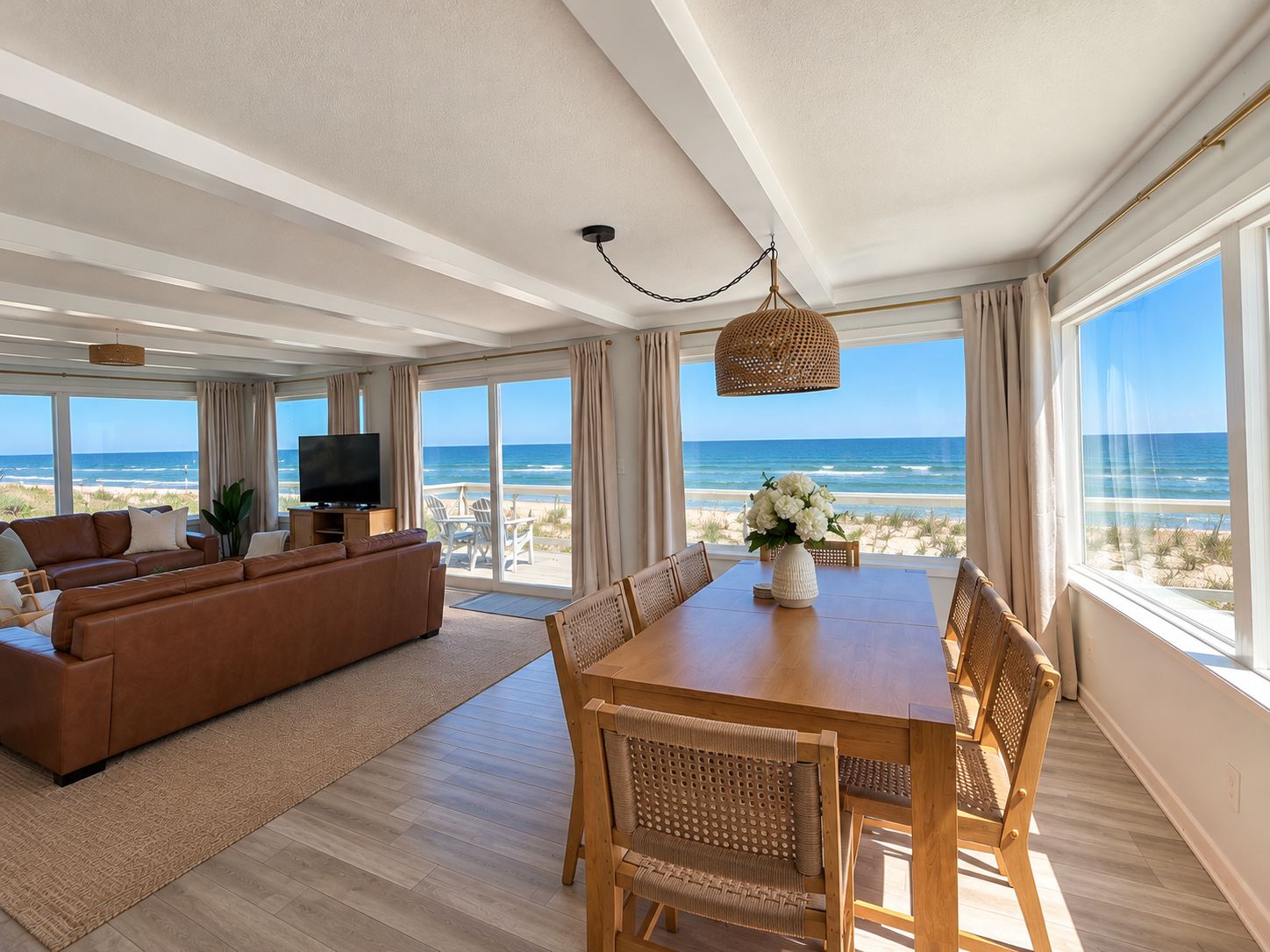 A spacious living room with a large sofa, a dining table with chairs, a TV, and a view of the beach through large windows.