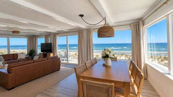 A spacious living room with a large sofa, a dining table with chairs, a TV, and a view of the beach through large windows.