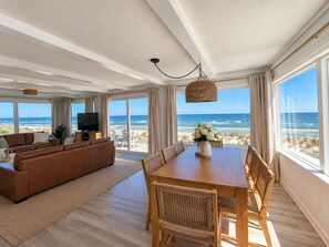 Een ruime woonkamer met een grote bank, een eettafel met stoelen, een televisie en uitzicht op het strand door grote ramen.