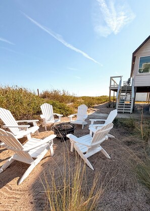 A beachside outdoor seating area with Adirondack chairs arranged around a fire pit, a wooden deck, and a building with a large window and a staircase. - Oceanfront Escape, Grill, Hammocks, Fire Pit, & Stunning Views (Ponte Vedra Beach)