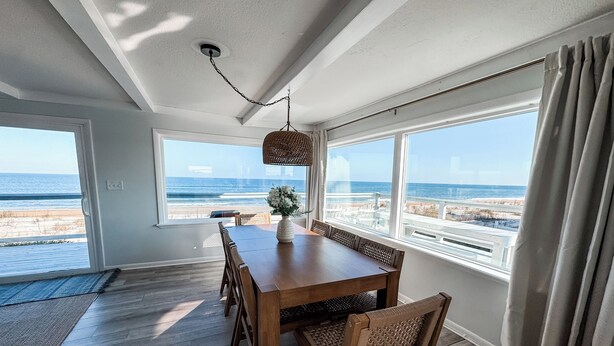 A dining area with a wooden table and chairs, large windows with a beach view, and a hanging light fixture. - Oceanfront Escape, Grill, Hammocks, Fire Pit, & Stunning Views (Ponte Vedra Beach)