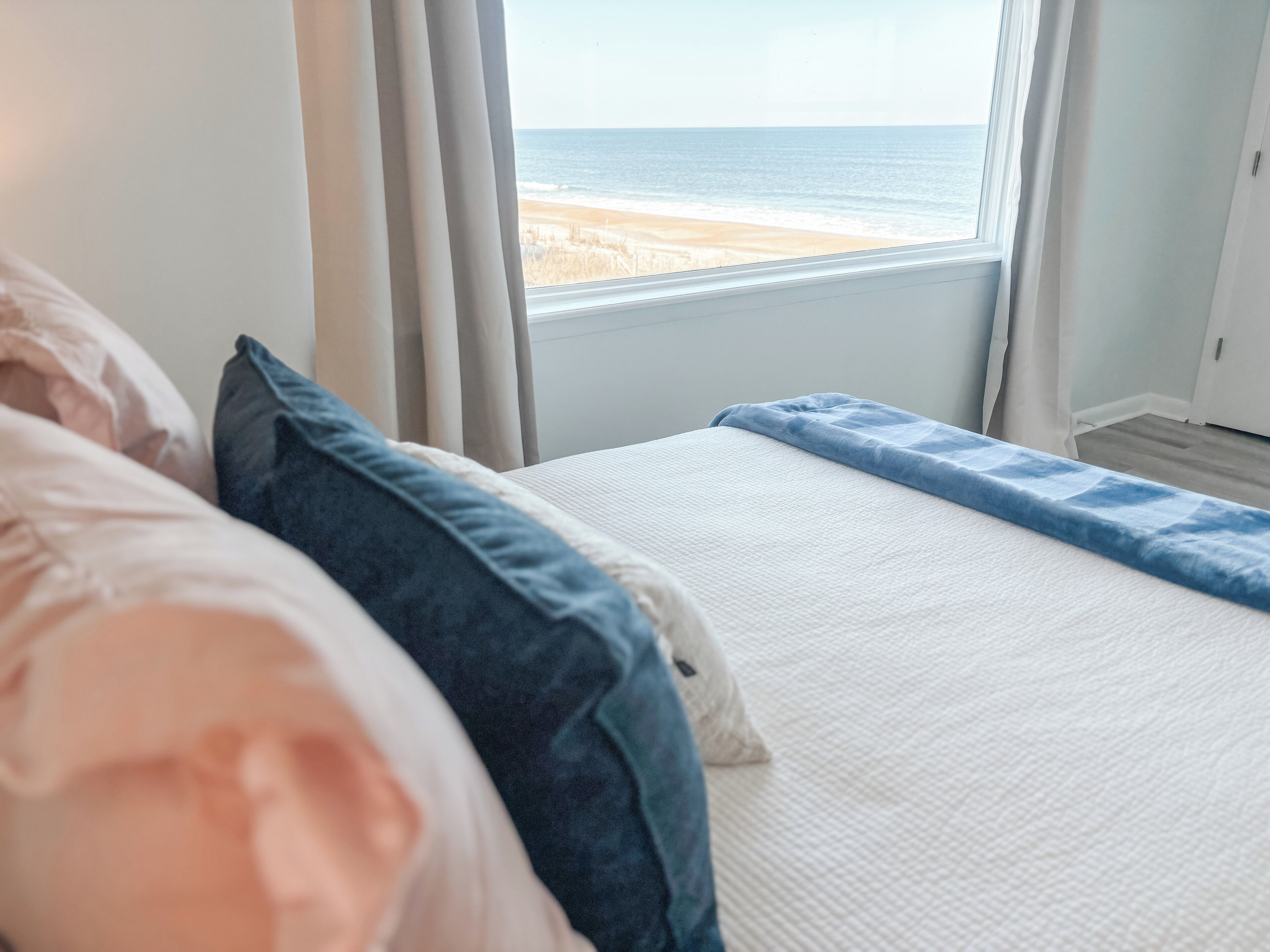 A bedroom with a bed, a view of the beach, and a window with curtains.