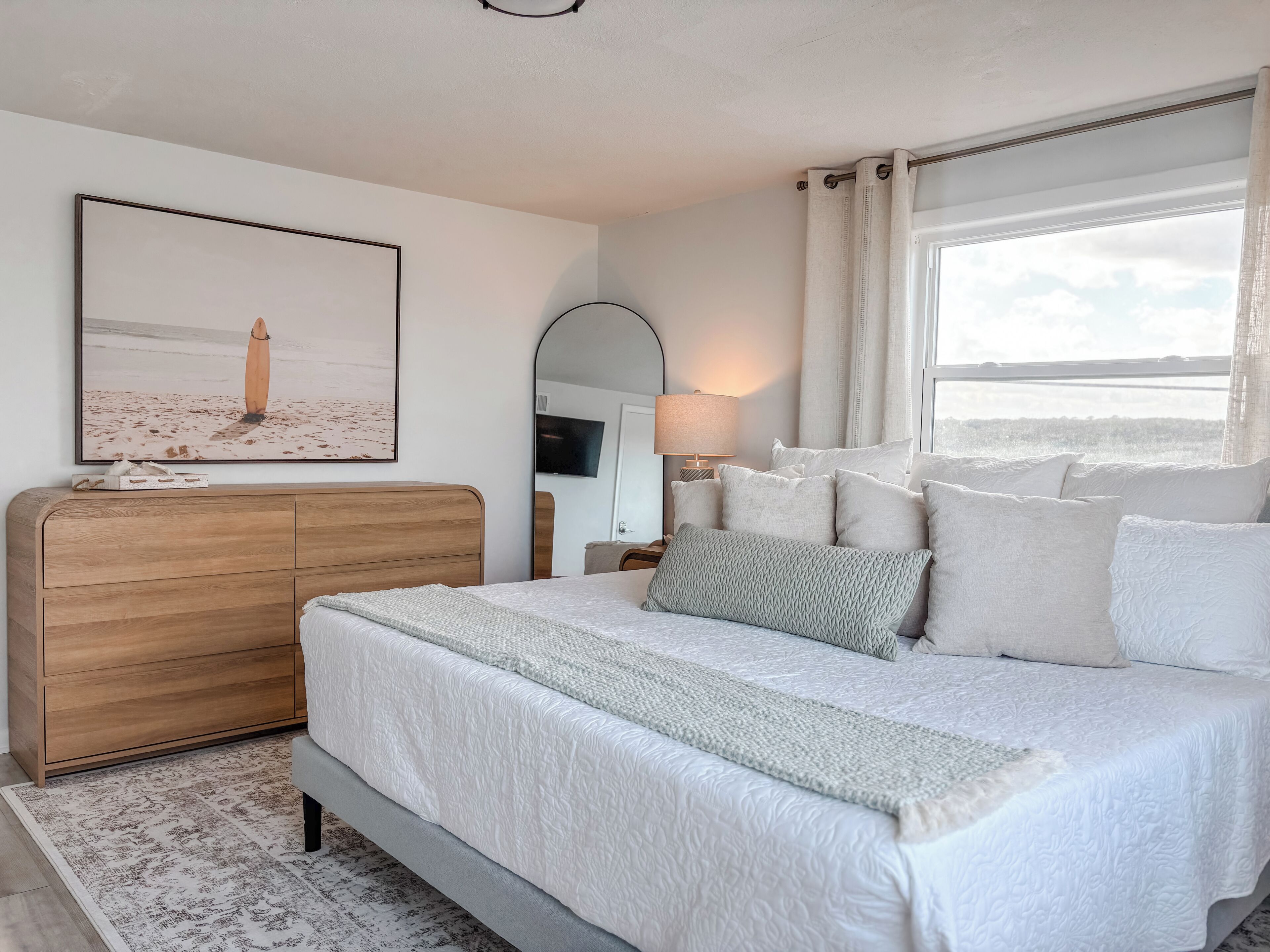 A neatly arranged bedroom with a bed, a wooden dresser, a TV, and a window with curtains.