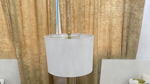 Habitación de hotel con mesita de noche blanca, lámpara dorada, cama blanca con ropa de cama floral y cortina con estampado dorado.