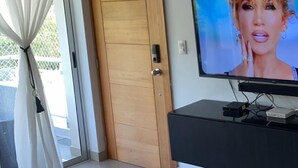 Una habitación con puerta de madera, un televisor sobre un soporte negro y vistas a través de un ventanal.