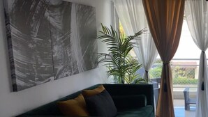 Un salón con un sofá verde, un cuadro abstracto grande, una planta en maceta y vistas al exterior a través de una ventana con cortinas translúcidas.