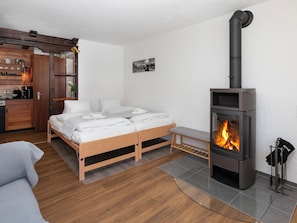Quarto com cama, um banco, piso de madeira, área de cozinha e lareira a lenha.