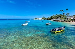 Helder turquoise water met boten, een zandstrand en een rij huizen met palmbomen.