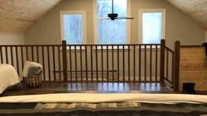 Une chambre avec un plafond en pente, des poutres en bois, un lit recouvert d’une couverture à motifs, une petite fenêtre et un ventilateur de plafond.