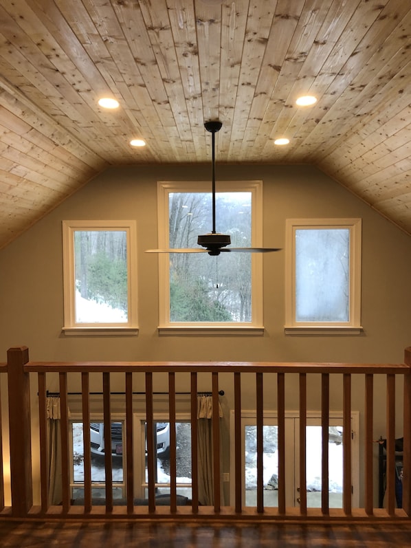 Una habitación con techo inclinado, vigas de madera, ventilador de techo y tres ventanas.