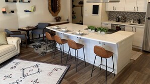 Una cuina moderna amb una illa central, tamborets d'esbarjo i una zona de menjador amb sofà i cadires.