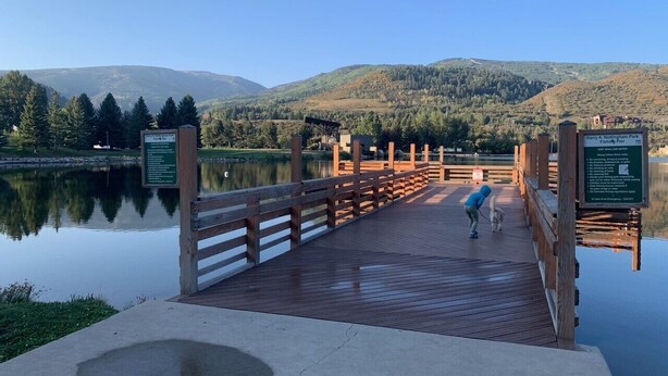Un passeig de fusta cap a un moll sobre un llac tranquil, envoltat de muntanyes i arbres.