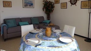 Ein Essbereich mit einem runden Tisch, gedeckt für vier Personen, einem grauen Sofa mit türkisfarbenen Kissen und Wanddekoration im Seestil.