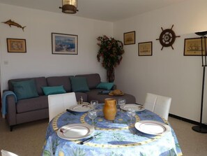 A dining area with a round table set for four, a grey sofa with teal cushions, and nautical-themed wall decor.