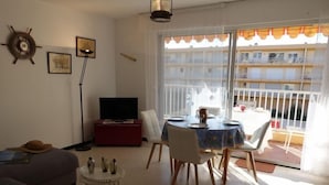 Ein Wohnzimmer mit Sofa, Fernseher auf einem Schrank, Esstisch mit Stühlen und einem Balkon mit Geländer.