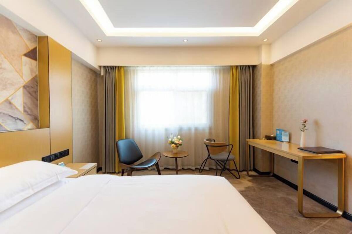 Une chambre d’hôtel avec un grand lit, un bureau, deux chaises, une petite table ornée de fleurs et une grande fenêtre avec des rideaux.