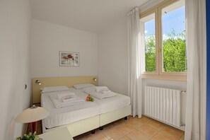 A clean, well-lit bedroom with a bed, a bedside table, a lamp, and a window with a view of greenery.