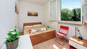 Ein Schlafzimmer mit Bett, Stuhl, Schreibtisch, Fenster und einer Pflanze.