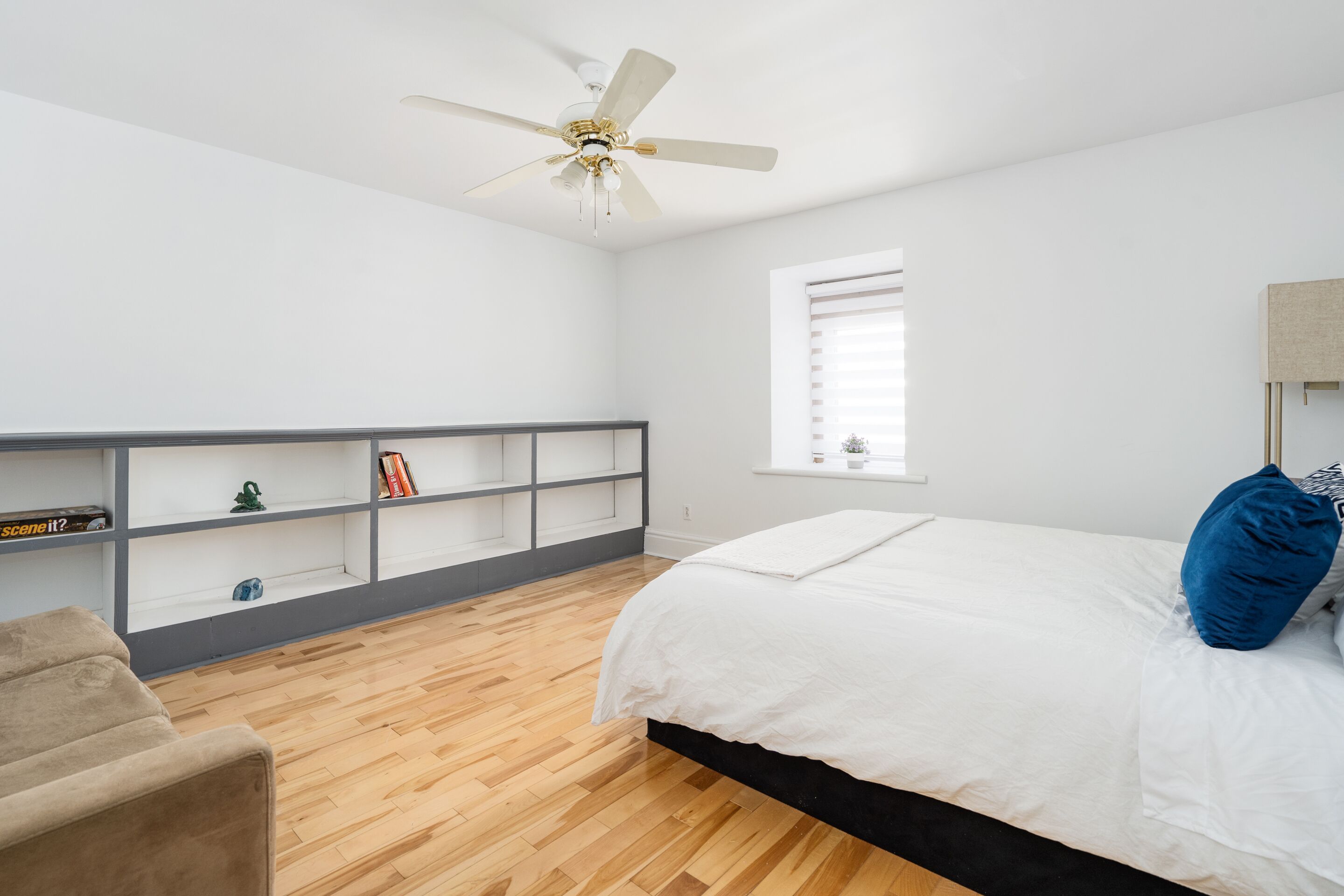 Une chambre à coucher avec un lit, un ventilateur de plafond, une fenêtre, une bibliothèque et un plancher en bois.
