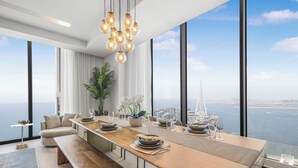 A modern dining area with a long wooden table set for a meal, large windows offering a city and water view, and a hanging light fixture. - EDEN'S Homes & Villas - 52/42 Emaar (Dubai)
