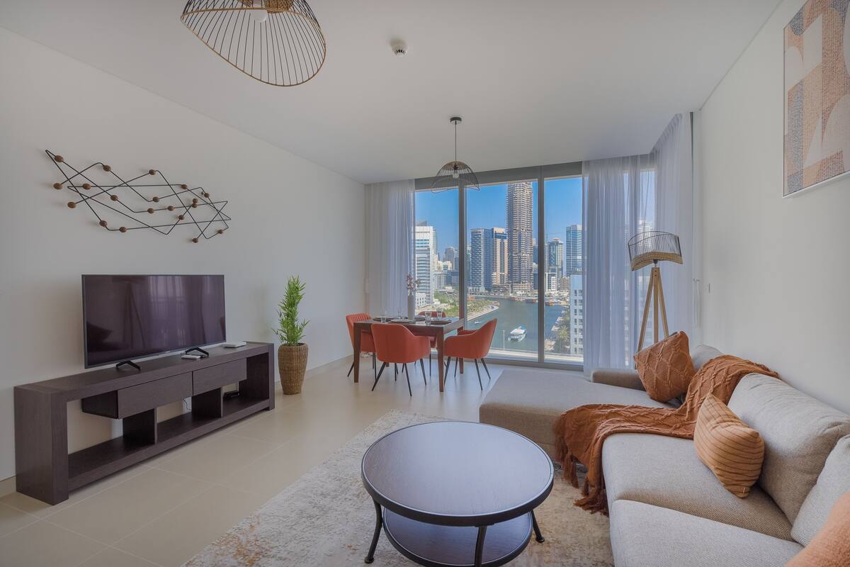 Een moderne woonkamer met een flatscreen televisie, een eethoek met oranje stoelen, een bank met een plaid en een groot raam met uitzicht over de stad.