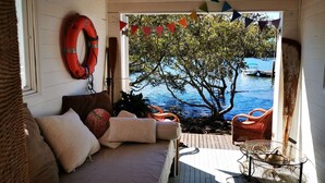 Et hyggeligt rum med træterrasse, sofa med puder, redningskranse på væggen og udsigt til vand og både ude.