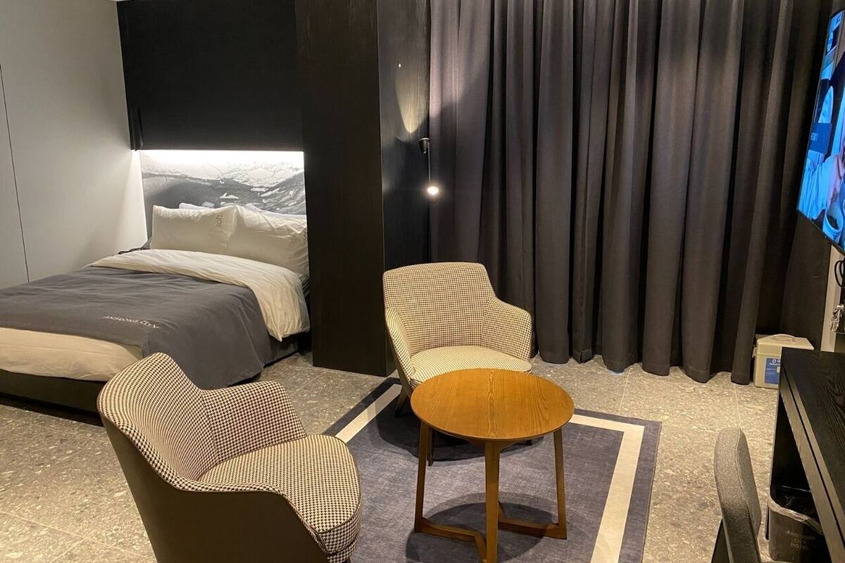 Habitación de hotel moderna con cama, un sillón a cuadros, una pequeña mesa redonda y un amplio ventanal con vistas a un paisaje nevado.