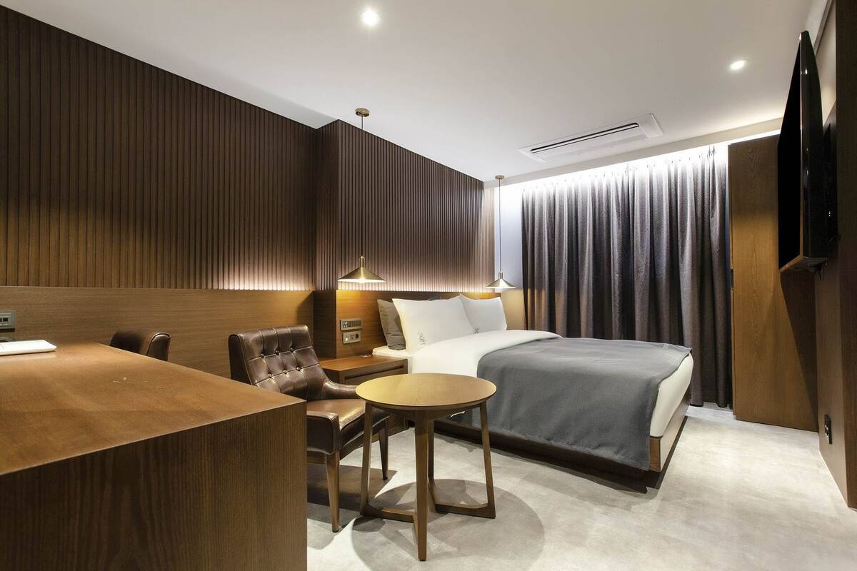 Habitación de hotel moderna con pared de paneles de madera, una cama con colcha gris, un escritorio con una computadora portátil, una silla de cuero, una mesita redonda pequeña y un televisor a la derecha.