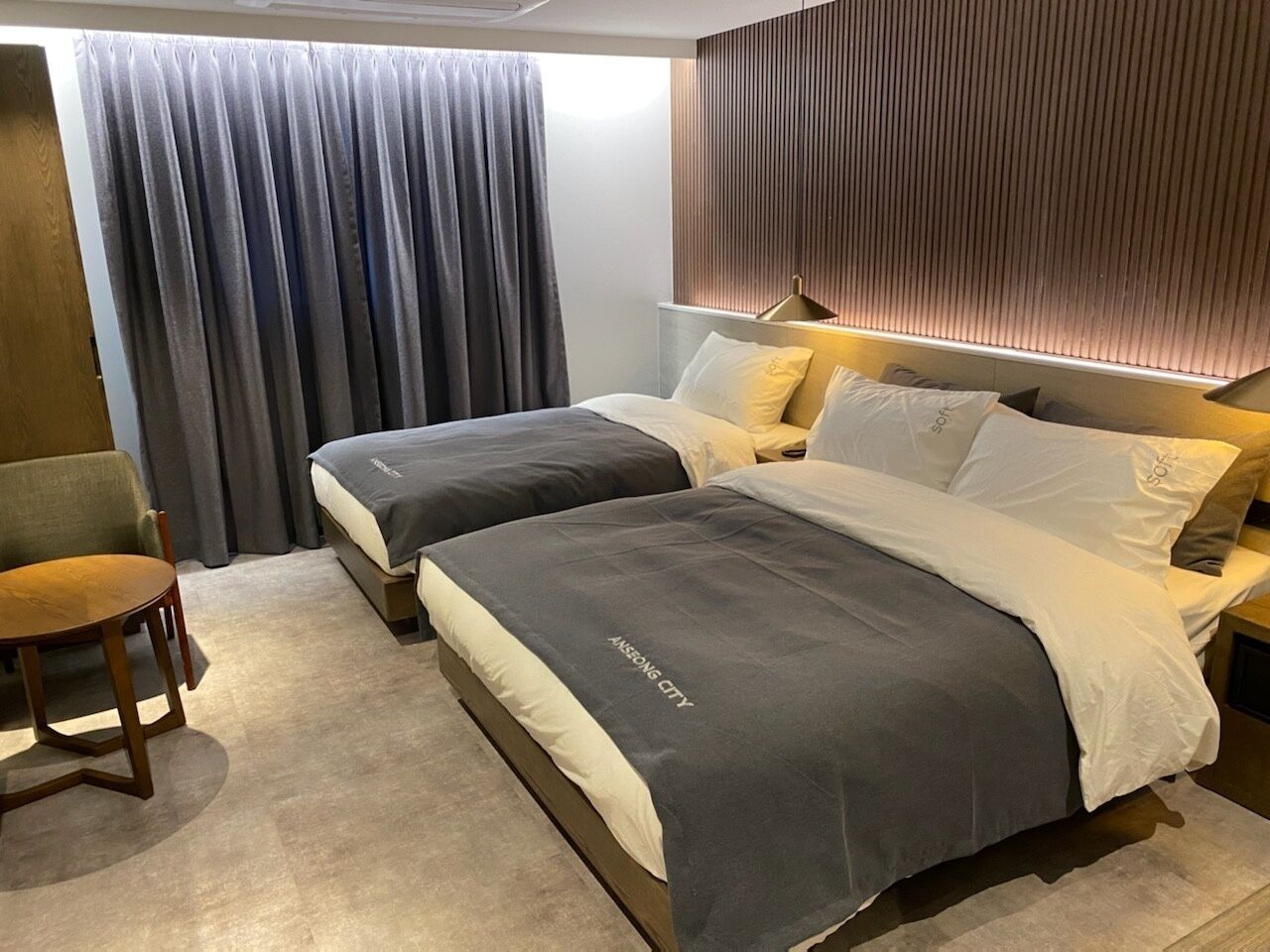 A hotel room with two beds, a wooden chair, a table, and a large window with curtains.