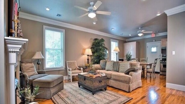 A spacious living room with a large sofa, ottoman, and a coffee table. There is a fireplace, a large window with blinds, and a ceiling fan. - Trailside Manor - 0.5mi from UofA Campus! (Fayetteville)