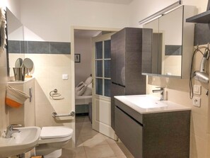 Ein modernes Badezimmer mit Waschbecken, Toilette und Waschtisch. Über dem Waschtisch hängt ein Spiegel und es gibt einen Handtuchhalter. Das Badezimmer hat eine Tür zu einem weiteren Raum.