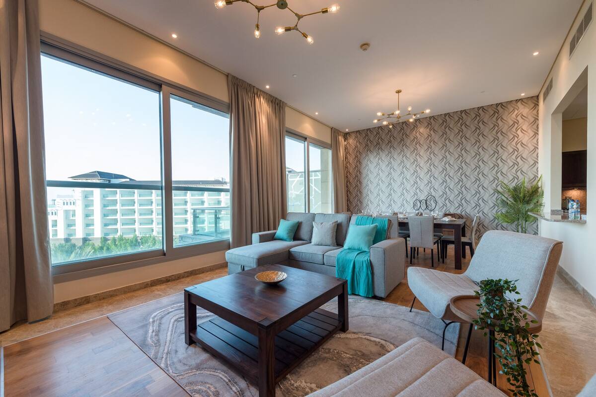 Una sala de estar moderna con sofá, sillón y mesa de centro, ventanas amplias con cortinas, un muro con diseño y una zona de comedor al fondo.