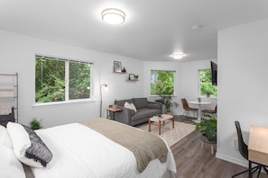 A modern bedroom with a large bed, a grey sofa, a small table, and a desk. There are large windows with views of greenery.