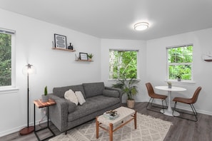 A living room with a grey sofa, a coffee table, a floor lamp, a small table with a plant, and two chairs. There are two windows with greenery outside.
