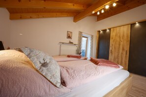A bedroom with a wooden ceiling, a large bed with pink bedding, a window with curtains, and a wardrobe.