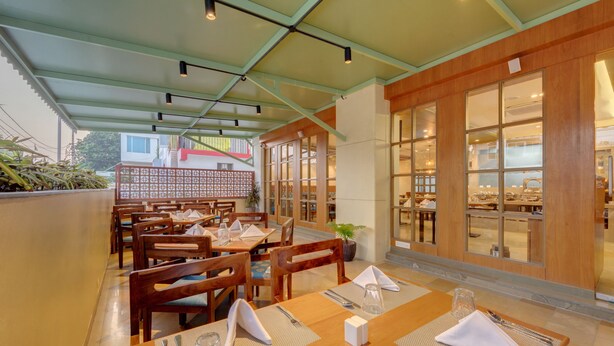 A restaurant with wooden tables and chairs, a green ceiling, and large windows. - The Beacon Hotel Vadodara (Vadodara)