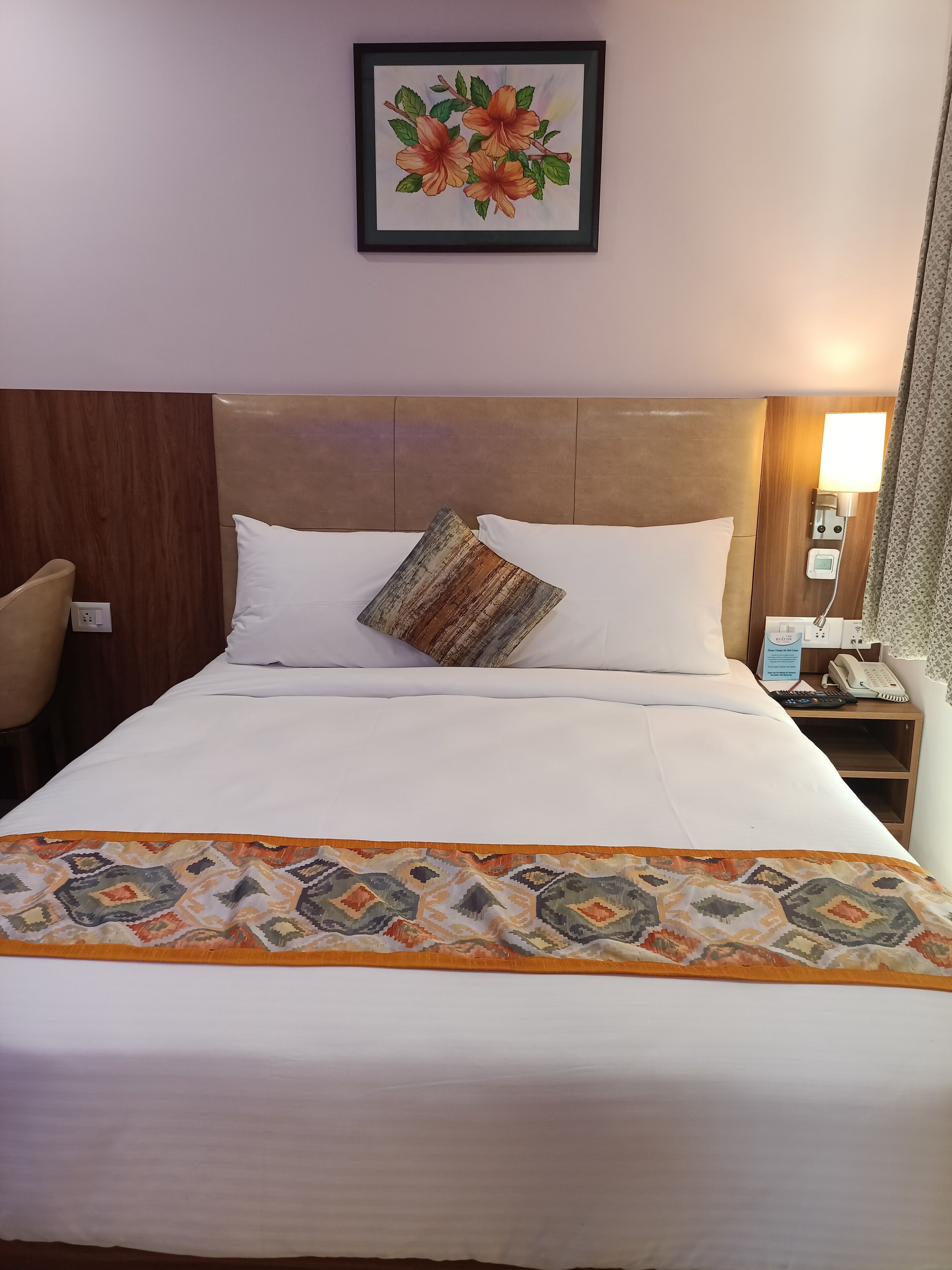 A hotel room with a bed, a nightstand with a phone and lamp, a chair, and a framed picture on the wall.