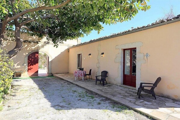 Une cour avec une table et des chaises, une porte rouge et un arbre qui offre de l’ombre.