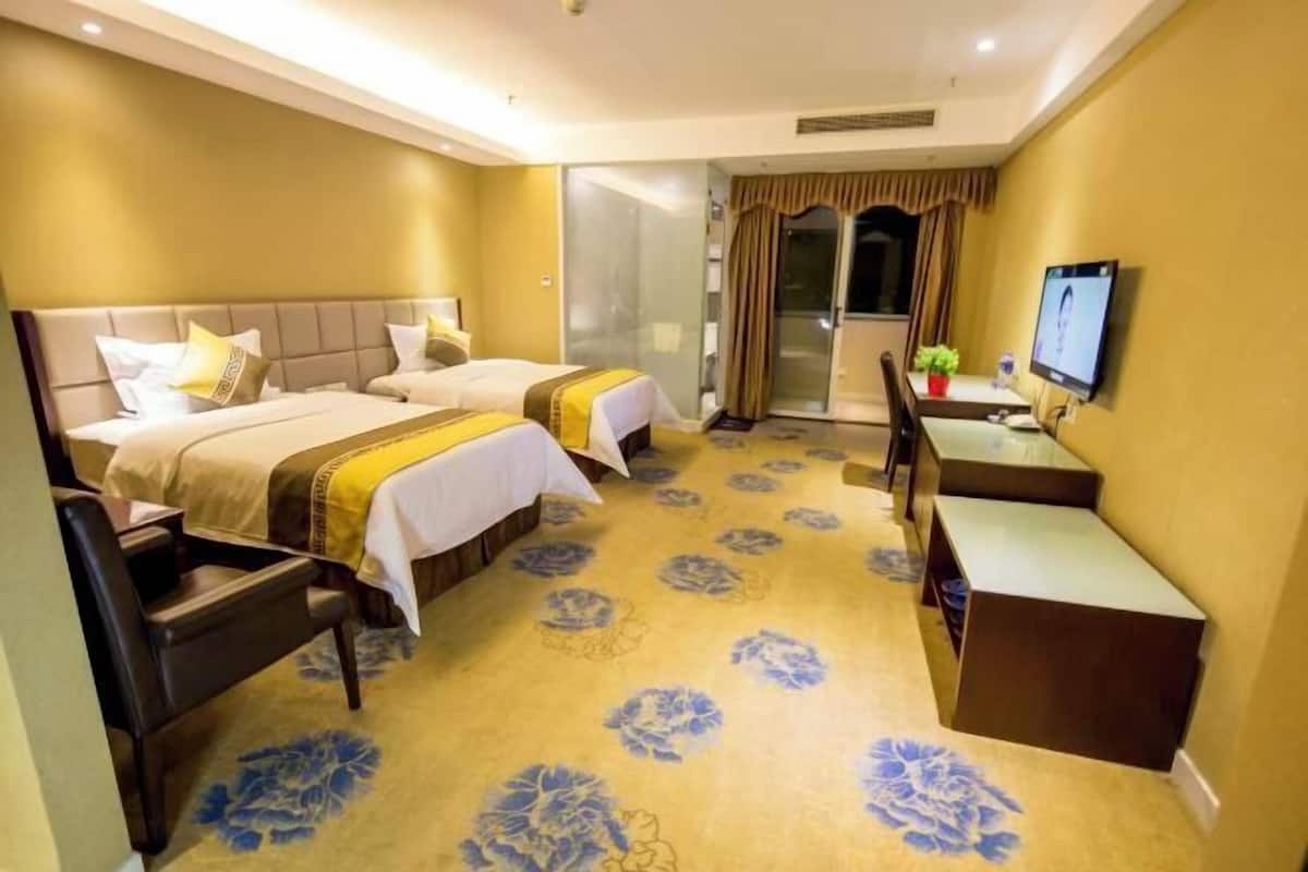 A hotel room with two beds, a desk, a chair, a TV, and a bathroom with a glass door.