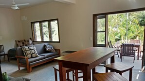 Una sala de estar con una mesa de comedor de madera, un sofá gris con cojines estampados y vistas a un jardín exuberante a través de amplios ventanales.
