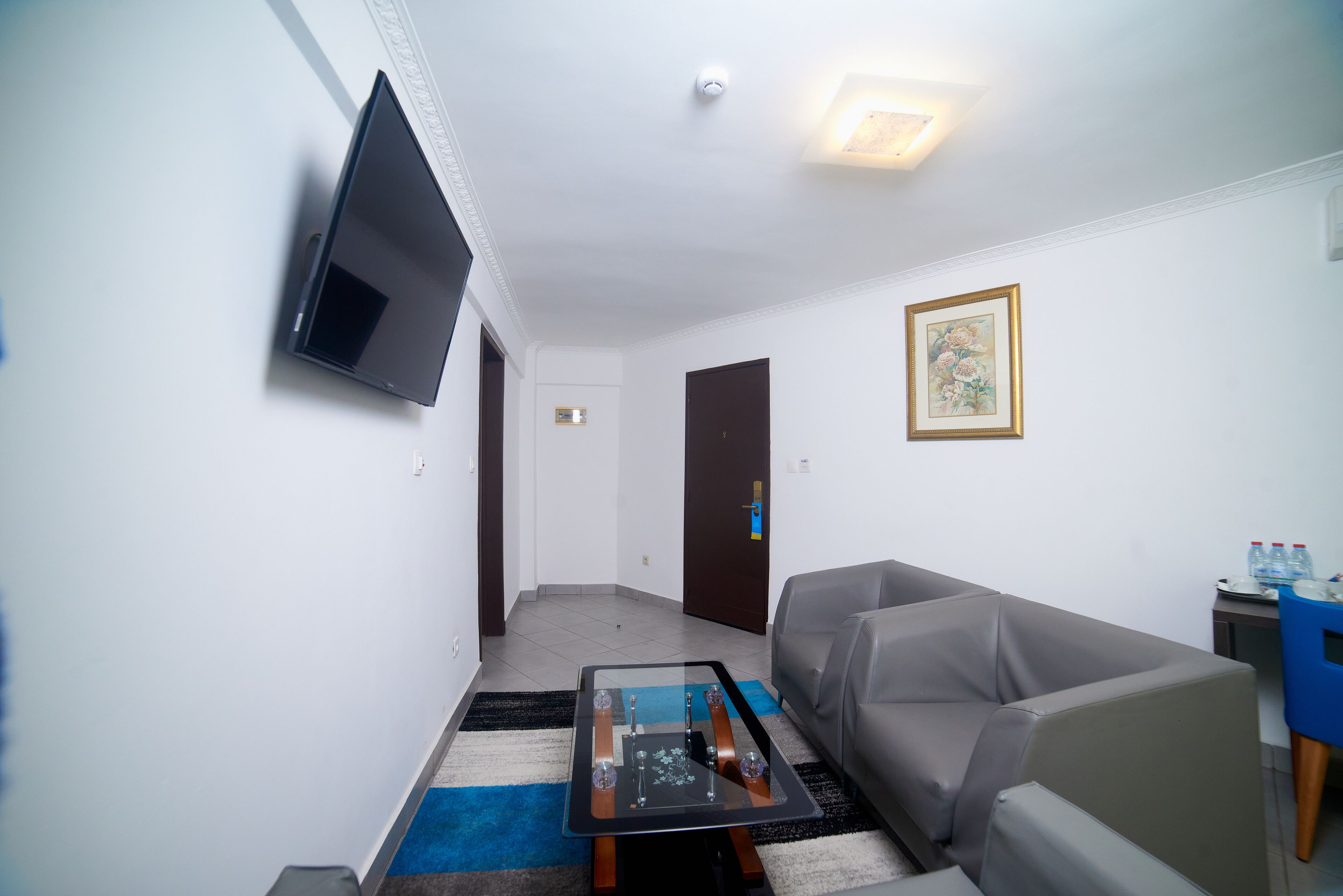 Een woonkamer met een flatscreen televisie aan de wand, een grijze bank, een salontafel met glazen blad, en een ingelijste foto aan de wand.