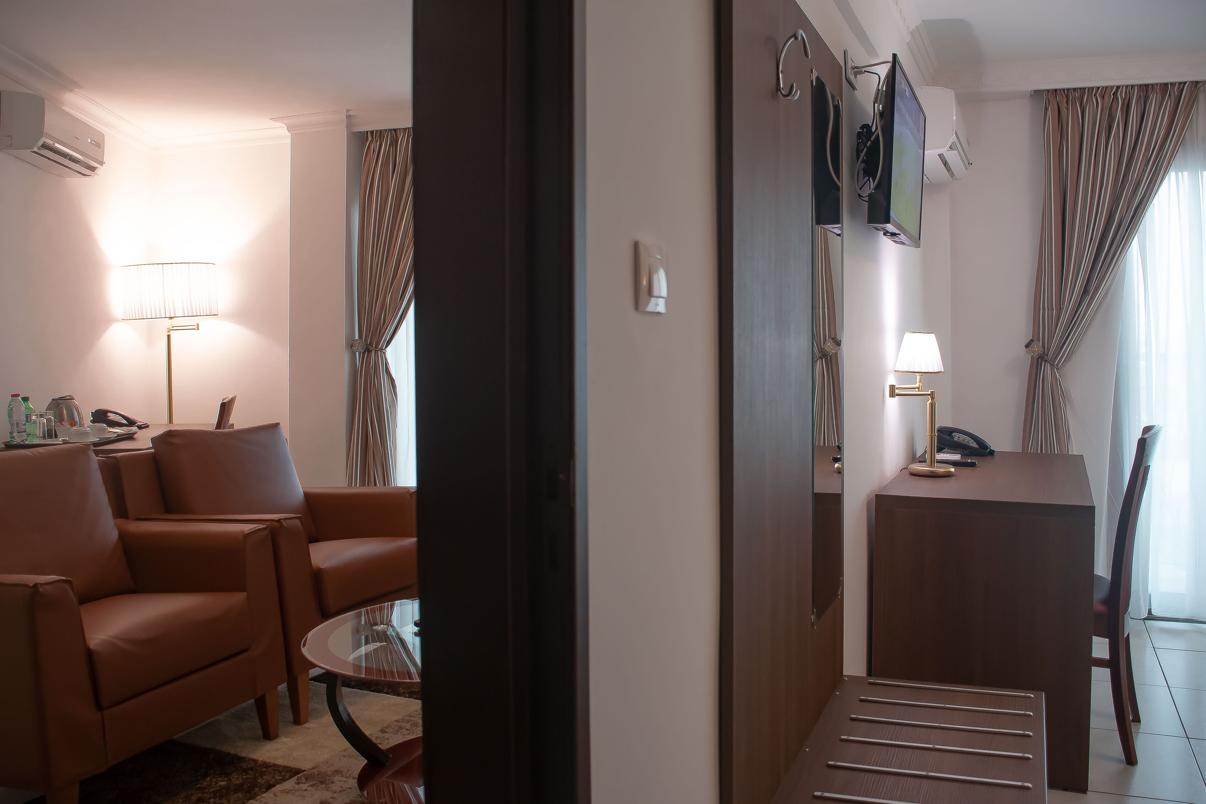 A hotel room with a brown leather sofa, a glass coffee table, a desk with a lamp, and a TV mounted on the wall.