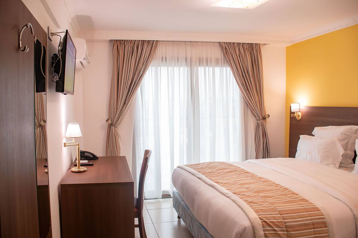 A hotel room with a bed, a desk with a lamp, a telephone, and a window with curtains.