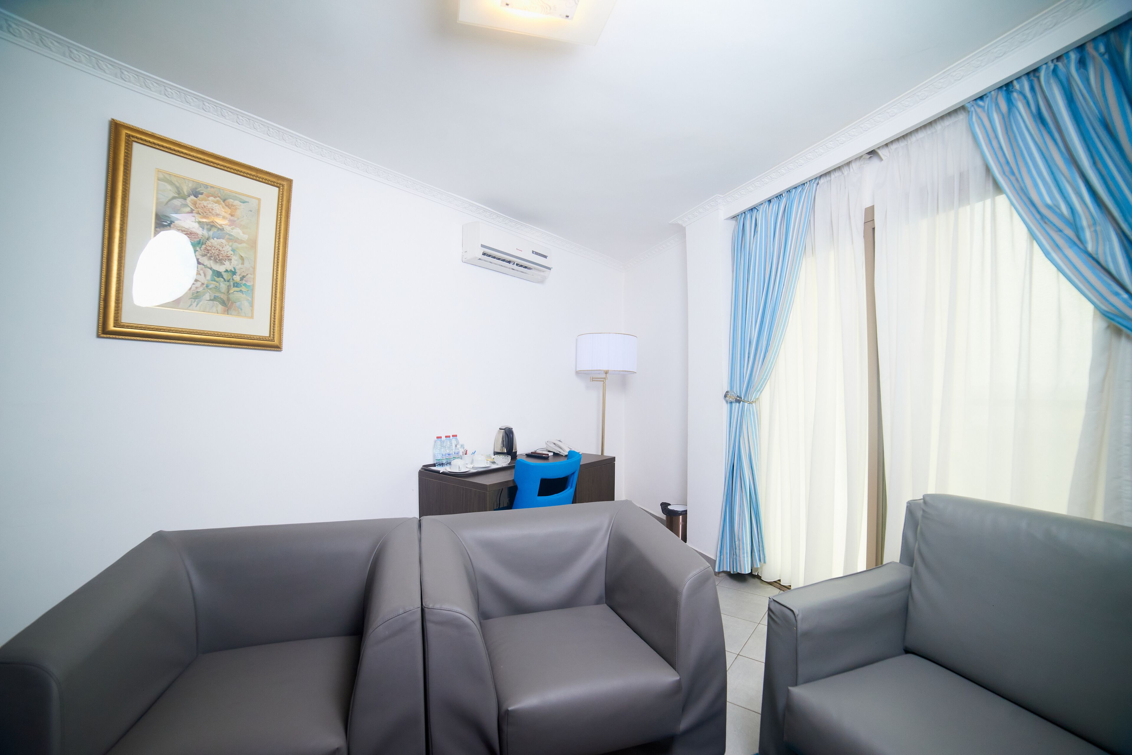 A hotel room with two grey sofas, a desk with a chair, a framed floral artwork, an air conditioning unit, and a window with blue curtains.