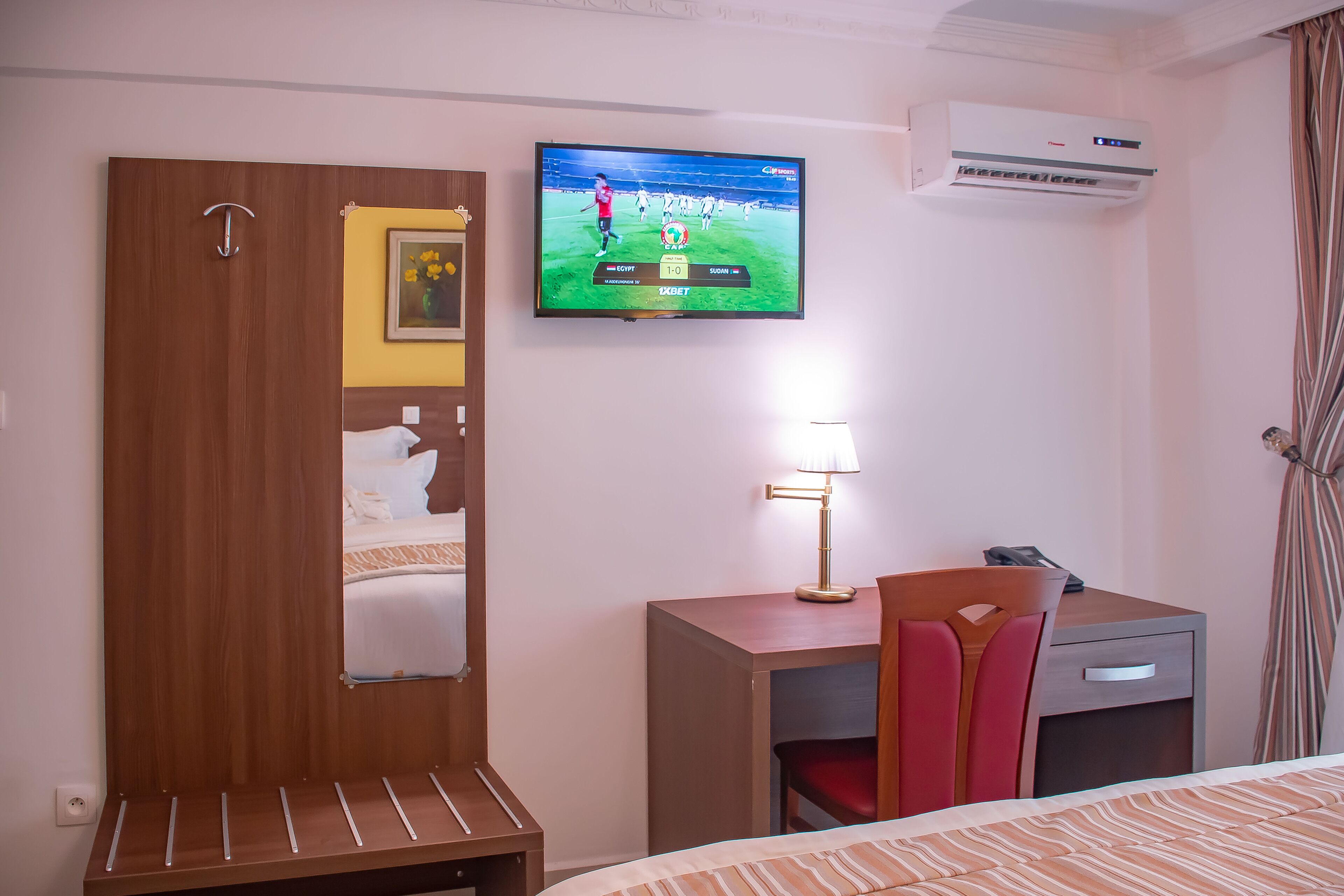 A hotel room with a bed, a desk with a chair, a television displaying a sports game, and an air conditioning unit.