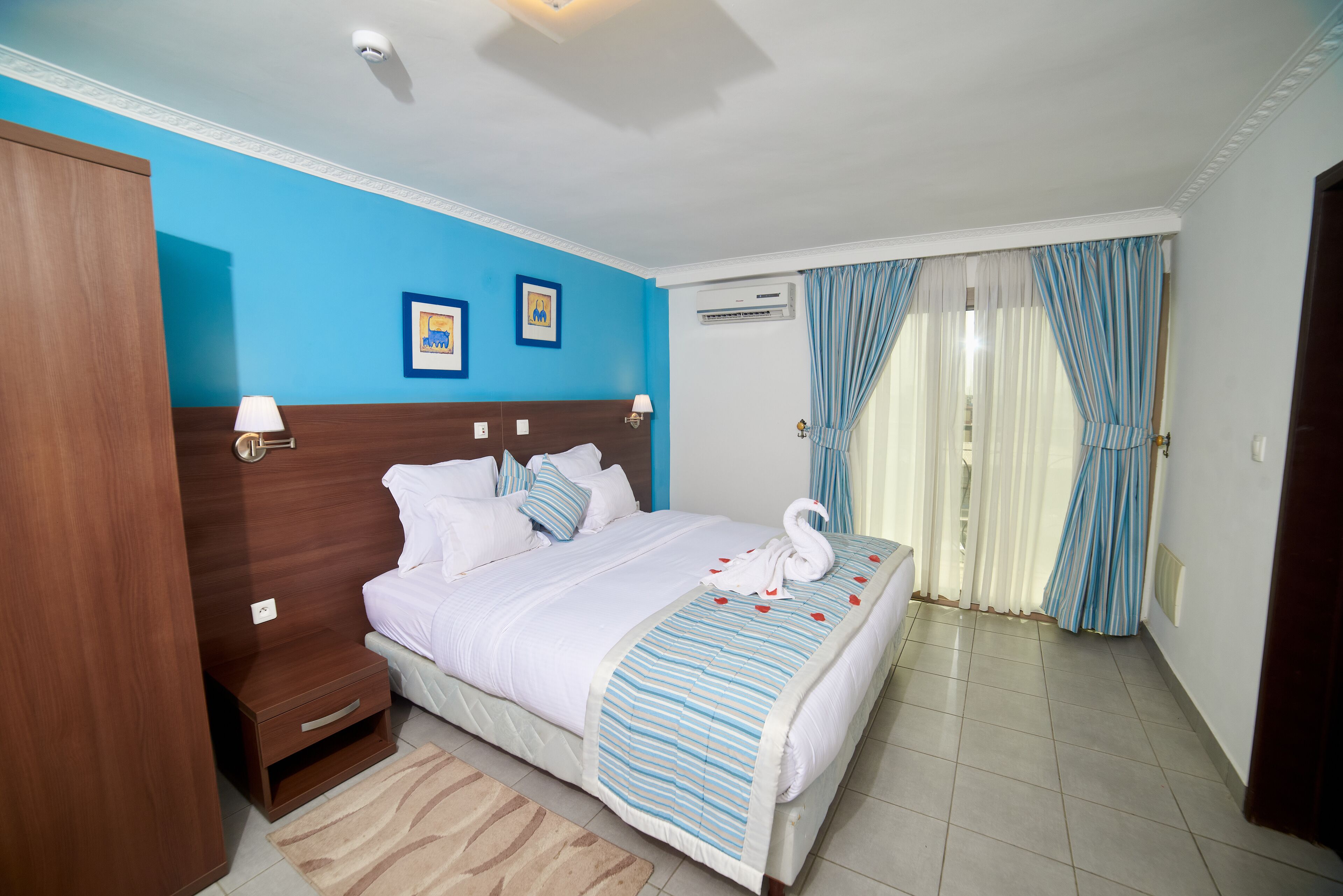 A hotel room with a bed, wooden headboard, bedside tables, a nightstand, a blue-striped bedspread, a white swan decoration, and a window with curtains.