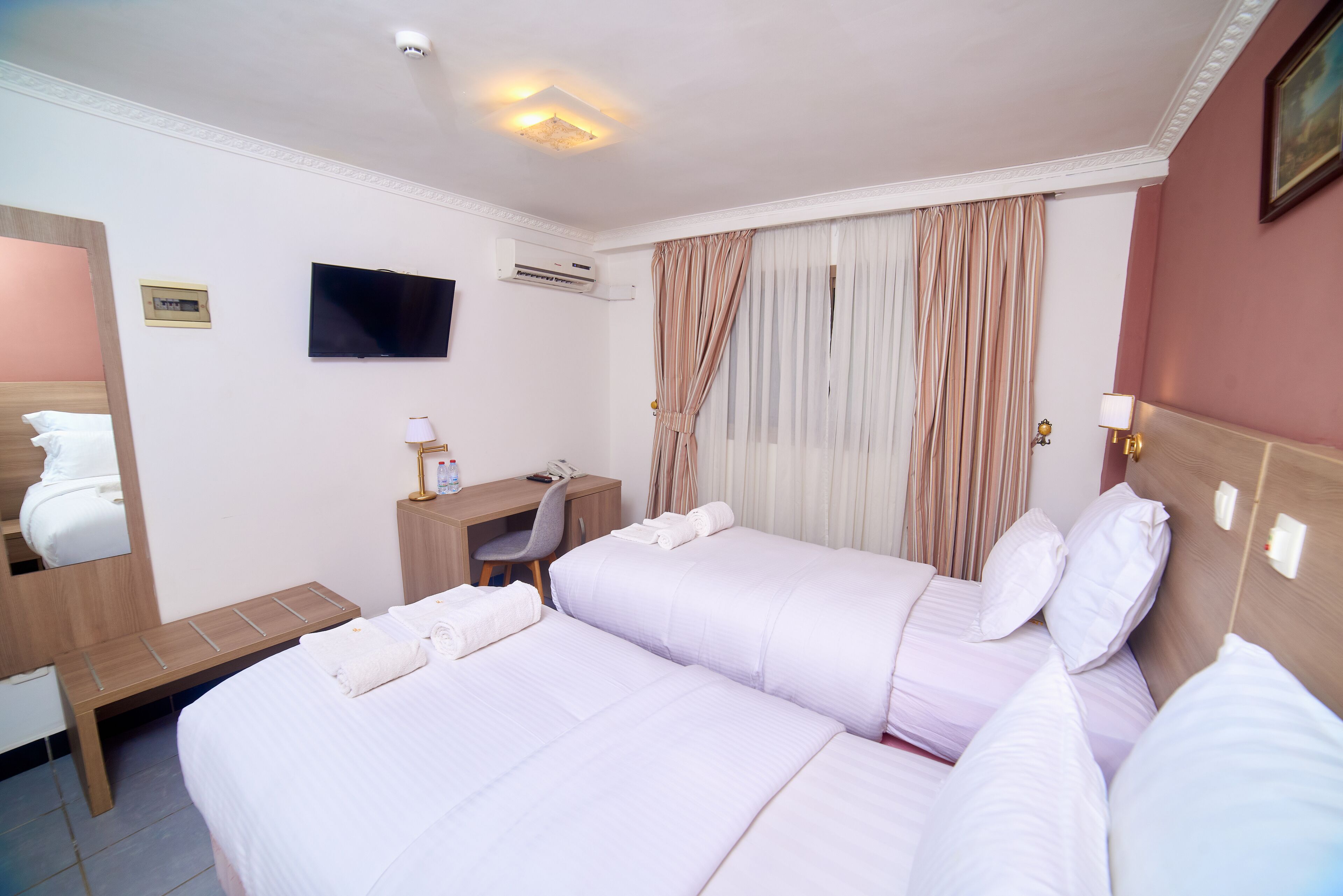 A hotel room with two beds, a desk, a chair, a TV, and an air conditioning unit.