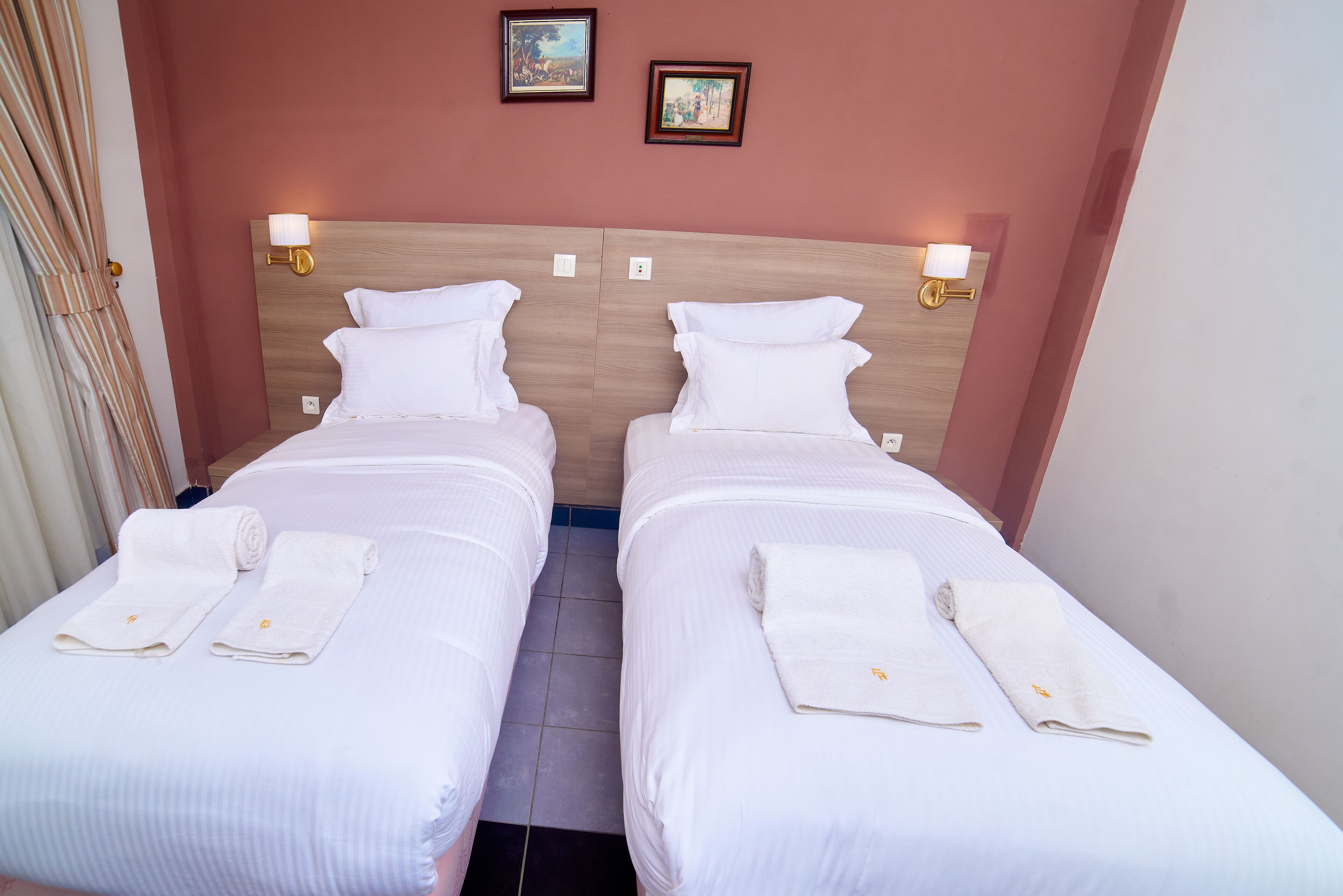 A hotel room with two single beds, each with white linens and towels, wooden headboards, and wall-mounted light fixtures.