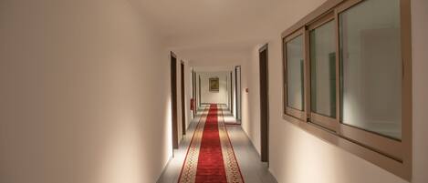 Un pasillo largo y estrecho con alfombra roja, paredes blancas y ventanas amplias a un lado.
