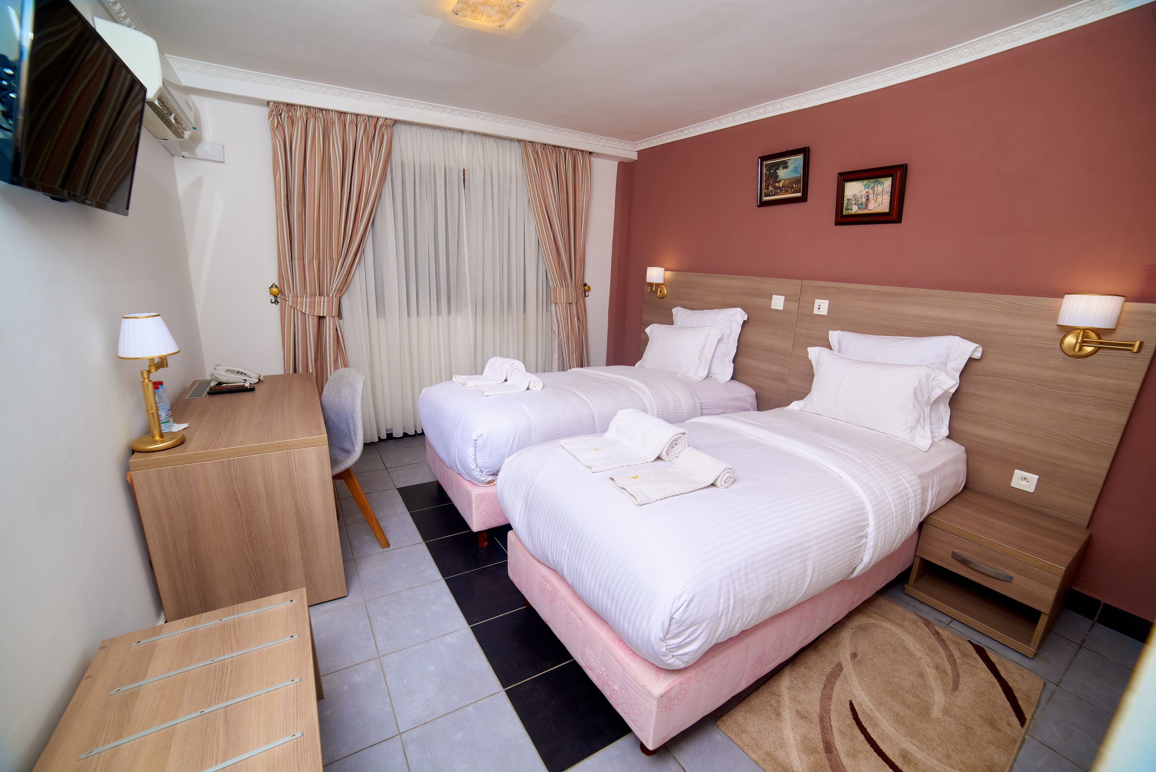 Quarto de hotel com duas camas, uma escrivaninha, uma cadeira, uma televisão e uma janela com cortinas.